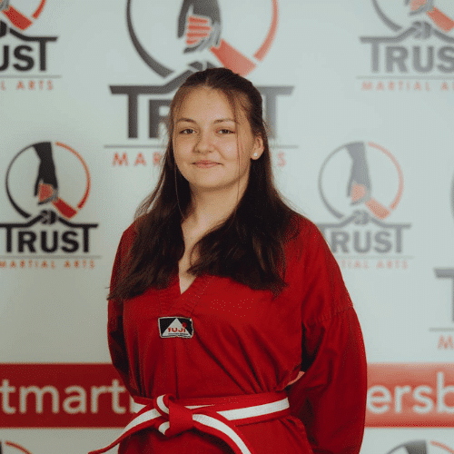 Junge Taekwondo-Schülerin der TRUST Martial Arts Schule Backnang im roten Dobok mit Rot-Weiß-Gurt vor dem TRUST-Hintergrund.