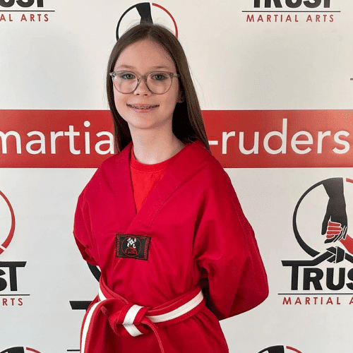 Junge Taekwondo-Schülerin der TRUST Martial Arts Schule Backnang im roten Dobok mit Rot-Weiß-Gurt und Brille vor dem TRUST-Hintergrund.
