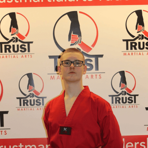 Taekwondo-Schüler der TRUST Martial Arts Schule Backnang im roten Dobok mit Brille vor dem TRUST-Hintergrund.